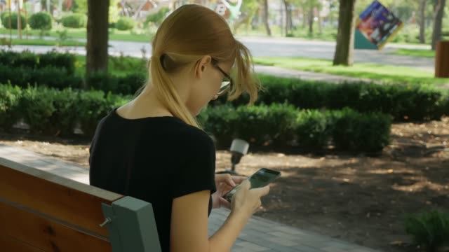 Feliz-chica-estudiante-usando-un-teléfono-inteligente-en-un-parque-de-la-ciudad-sentado-en-un-banco,-joven-mujer-sonriendo-uso-aplicación,-pelo-rubio-largo.