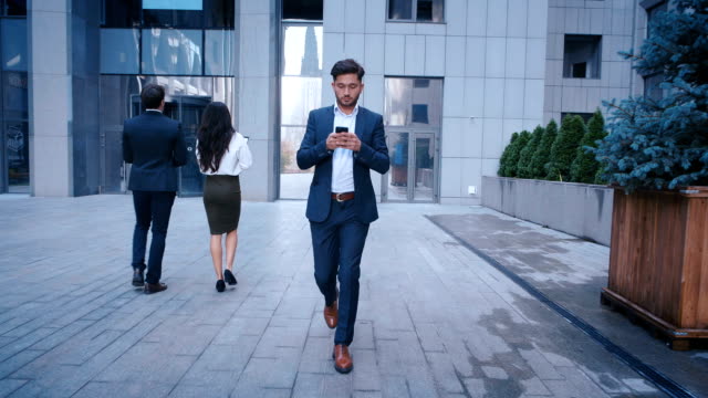 Young-Business-Man-Uses-Smartphone-While-Walking-on-the-Big-City-Business-District-Street.-Classically-Dressed.-Big-Office-Building-in-the-Background.-Looking-Successful,-Confident.