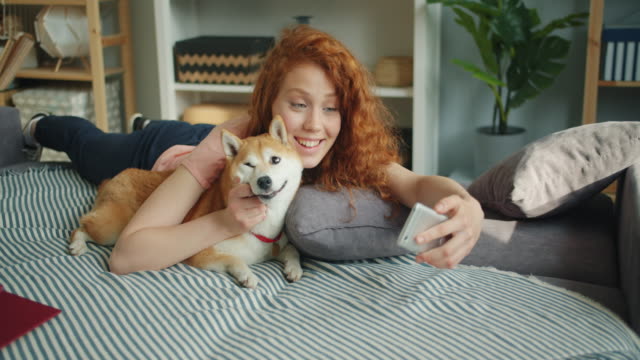 Loving-woman-kissing-cute-puppy-and-taking-selfie-with-smartphone-on-couch
