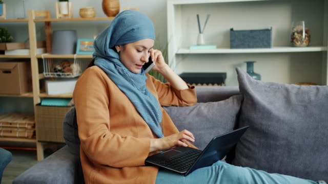 Cámara-lenta-de-la-empresaria-musulmana-hablando-por-teléfono-móvil-usando-computadora-portátil-en-casa