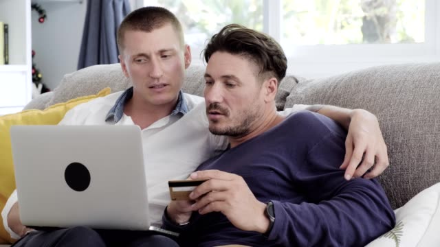 Pareja-gay-relajándose-en-el-sofá-usando-la-computadora-portátil.-Compras-en-línea.