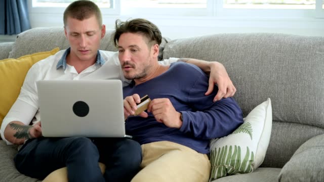 Gay-couple-relaxing-on-couch-using-laptop-computer.-Complete-purchase-and-kiss.