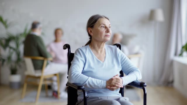 Tracking-shot-of-disabled-senior-woman-in-wheelchair-looking-away-thoughtfully,-then-turning-head-and-looking-at-camera-with-sadness-in-nursing-home,-other-aged-patients-in-background