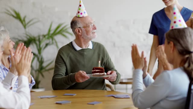 Älterer-Mann-spielen-Karten-mit-alten-Freunden-in-Party-Hüte,-während-Betreuer-bringt-ihm-Geburtstagstorte.-Glücklicher-Senior-Mann-immer-Wünsche-und-Umarmungen-und-Blasen-Kerze-aus-im-Pflegeheim