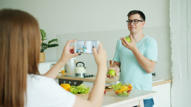 Student-jongliert-Äpfel-in-der-Küche,-wenn-Freundin-fotografieren-mit-smartphone