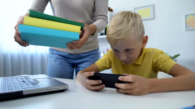 Mama-legt-Bücher-auf-den-Tisch,-freche-Junge-spielen-Smartphone-statt-Hausaufgaben