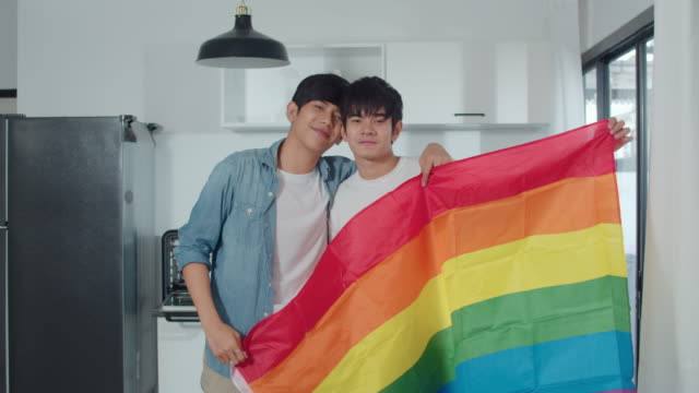 Porträt-Junge-asiatische-Homosexuell-Paar-Gefühl-glücklich-zeigt-Regenbogen-Flagge-zu-Hause.-Asia-LGBTQ+-Männer-entspannen-zu-schüchtern-Lächeln-blickend-in-die-Kamera,-während-Umarmung-in-der-modernen-Küche-zu-Hause-im-Morgen-Konzept.