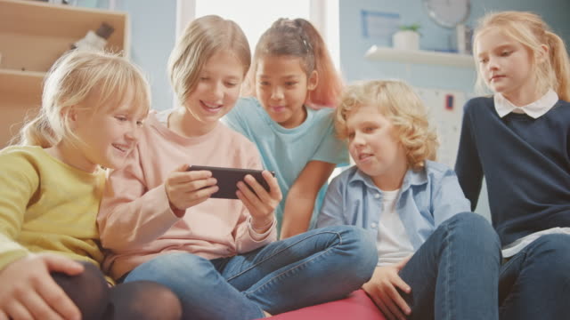 Diverso-grupo-de-niños-pequeños-lindos-sentados-juntos-en-las-bolsas-de-frijoles-usar-teléfono-inteligente-y-hablar,-divertirse.-Niños-navegando-por-Internet-y-jugando-videojuegos-en-línea-en-el-teléfono-móvil,-viendo-videos