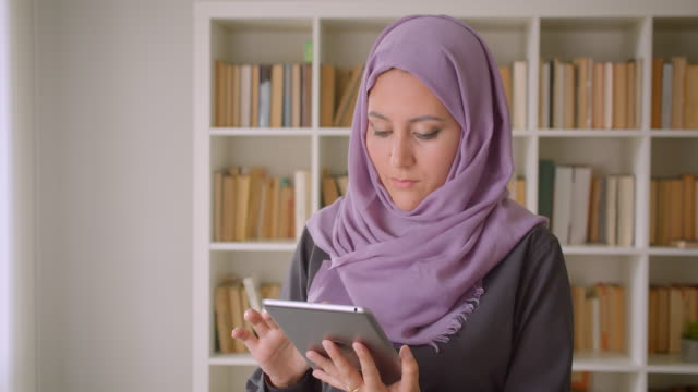 Retrato-de-primer-plano-de-joven-mujer-musulmana-en-hiyab-usando-la-tableta-y-mirando-la-cámara-de-pie-en-la-biblioteca