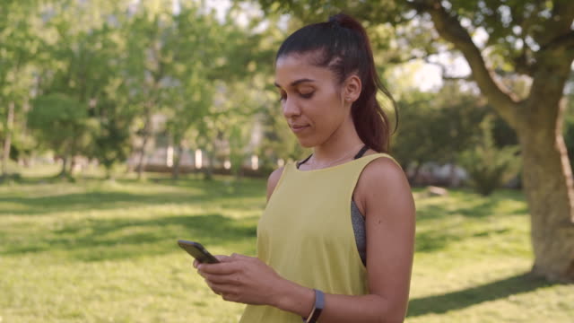 Porträt-einer-schönen-fit-lächelnden-jungen-Frau-in-Sportkleidung-stehend-im-Park-SMS-Nachrichten-auf-dem-Smartphone