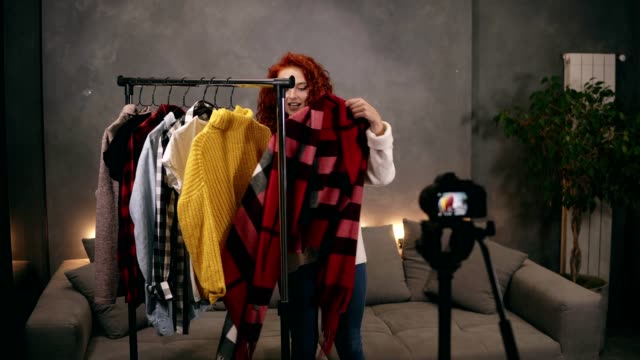 Stylish-red-headed-young-woman-vlogger-is-recording-video-for-online-blog-about-trendy-clothes-standing-nextto-the-rack-with-colourful-clothes-talking-and-gesturing.-Vlogging,-people-and-house-concept