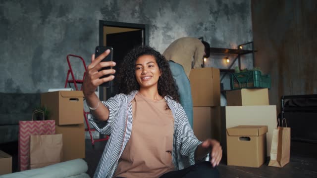 Multiracial-friends-moving-into-new-house.-Guy-unpacking-indoor-plants.-Girl-smiling,-waving-hand-and-recording-video-for-her-blog-using-smartphone