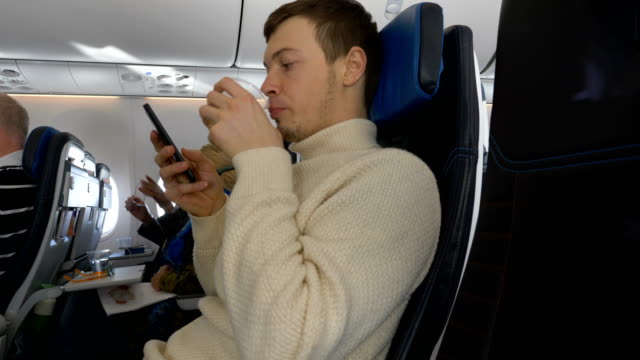 junger-Mann-schaut-auf-telefon,-während-im-Flugzeug-sitzt-trinkt-Wasser