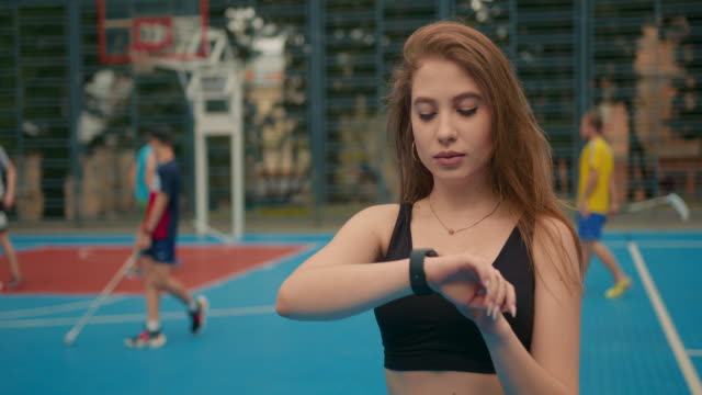 Close-up-shooting.-Portrait-of-a-sports-girl.-The-girl-is-looking-at-the-training-information-on-her-smartwatch.-People-are-playing-floorball-in-the-background.-4K