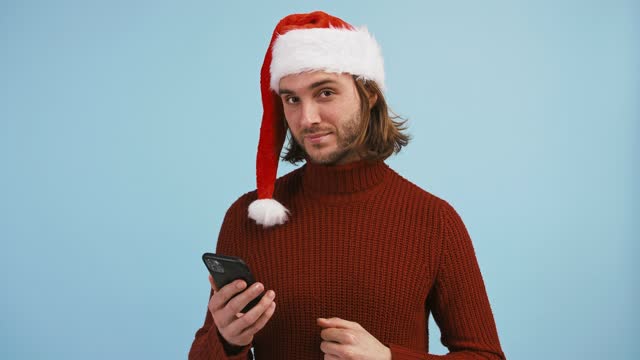 Young-male-in-Santa-hat-is-holding-smartphone-and-using-it,-smiling-and-showing-thumb-up,-posing-on-blue-background