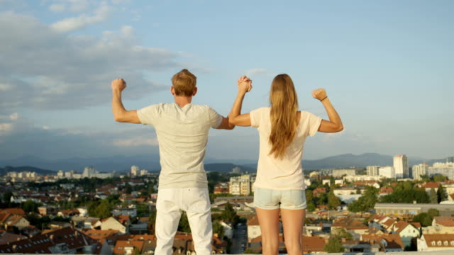 CLOSE-UP:-Girl-and-boy-standing-on-rooftop-above-the-city,-raising-hands-in-air