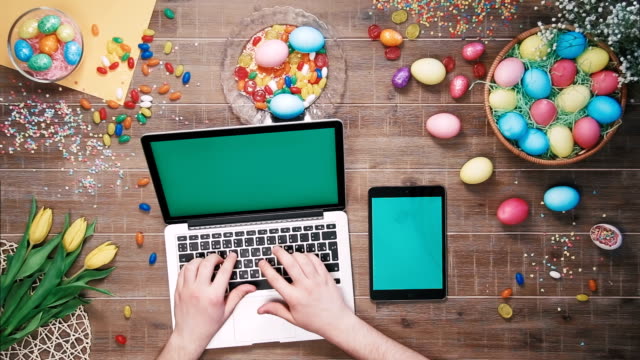 Hombre-con-ayuda-de-ordenador-portátil-y-tableta-digital-con-pantalla-verde-en-mesa-decorada-con-vista-superior-de-huevos-de-Pascua