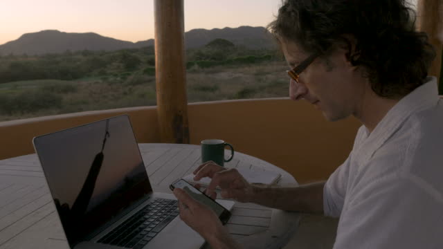 Man-scrolling-through-smart-phone-at-home-office-by-ocean-looking-at-email