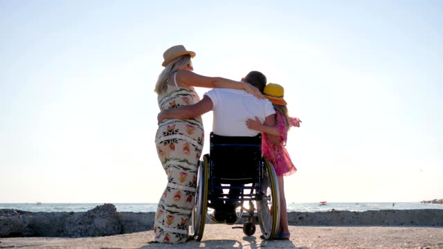 invalid-people-in-backlight,-daddy-in-wheelchair-hugging-daughter-on-background-blue-sky,-disabled-man