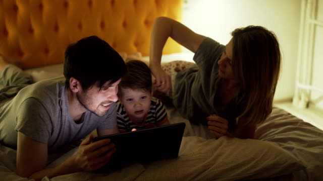 Glückliche-Familie-mit-kleinen-Sohn-liegend-im-Bett-zu-Hause-und-Surfen-social-Media-auf-dem-Tablet-PC-vor-dem-schlafen