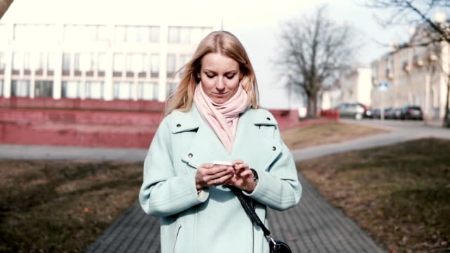Glückliche-kaukasischen-30er-Jahre-Frau-Tippen-auf-Smartphone.-Ziemlich-stylish-blonde-SMS-während-des-Gehens-in-der-Stadt.-Slow-motion