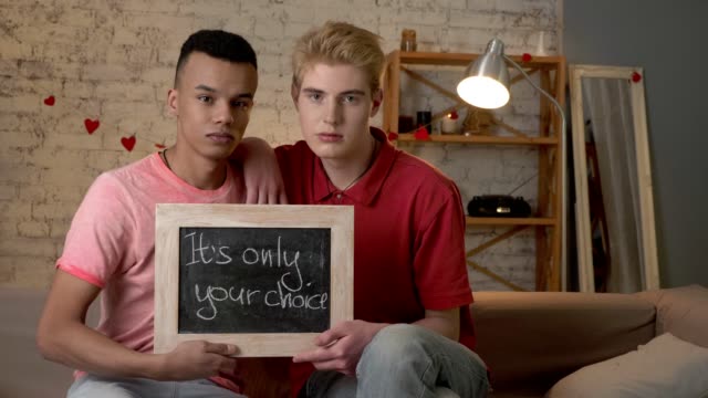 A-sad-international-gay-couple-is-sitting-on-the-couch-and-holding-a-sign.-It's-only-your-choice.-Look-at-the-camera.-Home-comfort-on-the-background.-60-fps