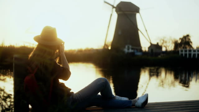 Female-photographer-sits-on-a-sunset-lake-pier.-Girl-with-camera-takes-a-photo-of-traditional-old-rustic-windmill.-4K