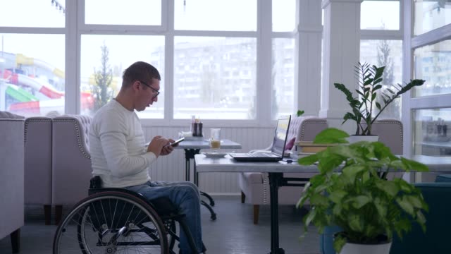 Freelance-business-man-in-a-wheelchair-wearing-glasses-uses-a-mobile-phone-and-working-on-laptop-sitting-at-a-table-in-a-cafe