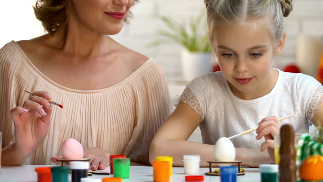 Mother-watching-daughter-painting-Easter-egg-for-first-time,-family-traditions
