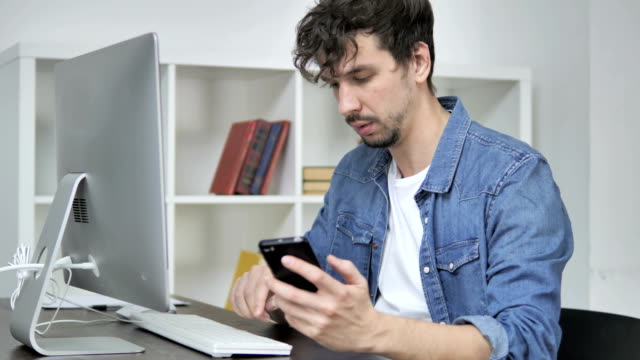Junge-Mann-mittels-Smartphone-während-der-Arbeit-am-Desktop
