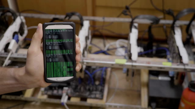 Closeup-of-it-specialist-hand-holding-smartphone-showing-mobile-app-for-mining-bitcoin-near-cryptocurrency-rig