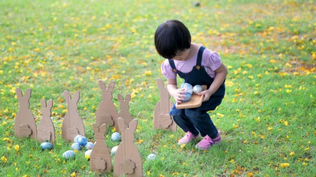 Cute-little-child-girl-on-Easter-day.-Girl-hunts-for-Easter-eggs-on-the-lawn-and-bunny-Made-of-paper-in-nature-or-park-and-sunlight.-Slow-Motion