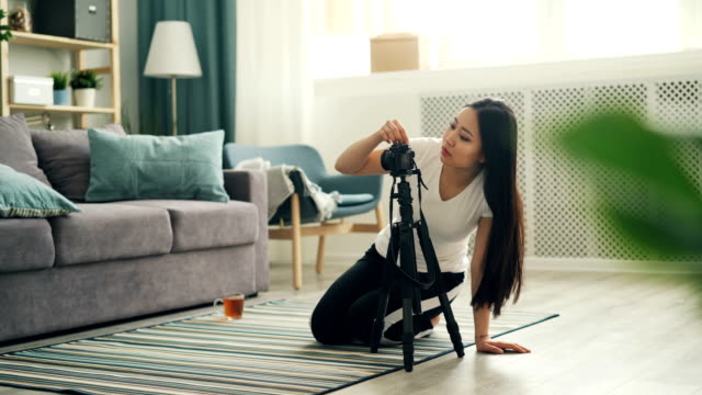 Atractivo-blogger-asiático-está-instalando-la-cámara-en-el-trípode-y-el-equipo-de-ajuste,-a-continuación,-sentado-en-el-suelo-y-grabación-de-vídeo-para-Vlog-en-línea.-Concepto-de-tecnología-y-estilo-de-vida.