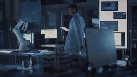 Professional-Heavy-Industry-Robotics-Engineer-Wearing-White-Coat-Holding-Tablet-Computer-Walks-Through-Laboratory.-In-the-Background-Facility-Full-of-Computers,-Various-Industrial-Design-Components-and-with-Robotic-Arm