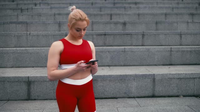 Plusgroße-blonde-gemischte-Rennracee-lächelnde-Frau-trägt-rote-Sportbekleidung-mit-Smartphone-im-Freien