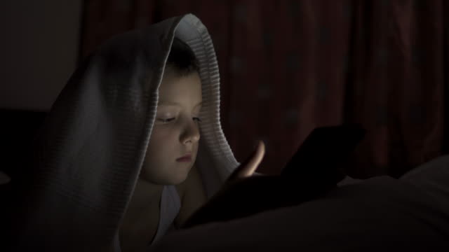 A-happy-boy-lies-in-bed-under-a-blanket-and-plays-on-a-tablet-in-a-game-in-the-dark.-The-face-of-the-child-is-lit-by-a-bright-monitor