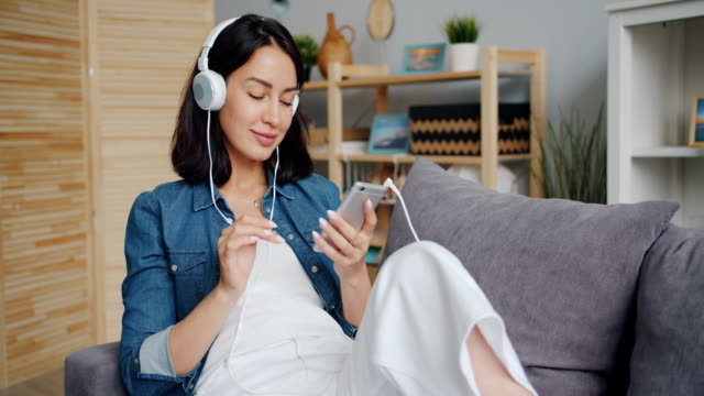 Female-student-enjoying-music-through-headphones-touching-smartphone-screen