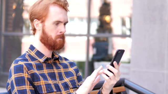 Barba-Joven-Hombre-usando-Smartphone-mientras-está-sentado-al-aire-libre