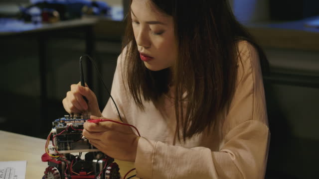 Young-asian-female-electronics-development-engineers-works-with-robot,-measuring-the-signal-in-the-electrical-circuits-of-robotics-prototype-in-workshop.-People-with-technology-or-innovation-concept.