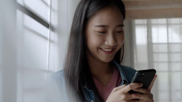 Portrait-smiling-young-asian-business-woman-holding-typing-mobile-phone-and-scrolls-through-social-media-feed-in-smartphone-standing-beside-window-at-home-office.