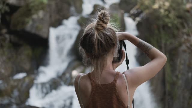 Ein-junges-Paparazzi-Mädchen-fotografiert-einen-schönen-großen-Wasserfall,-der-auf-Steinen-im-Dschungel-steht.-Rückansicht