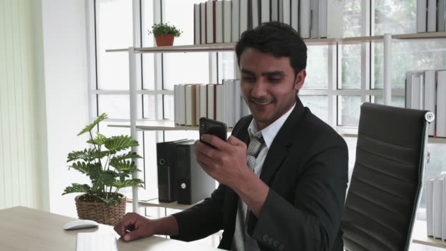 Hombre-de-negocios-feliz-en-traje-negro-usando-teléfono-inteligente-móvil-sentarse-en-el-escritorio-en-el-edificio-de-la-oficina.-Escribir-un-mensaje-en-su-smartphone.-Concepto-de-tecnología-de-comunicación-y-estilo-de-vida-empresarial.