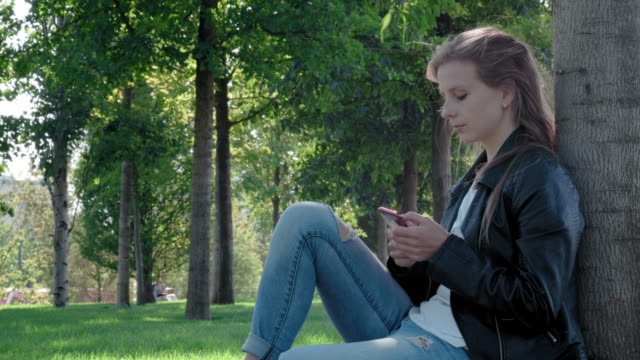 Young-beautiful-girl-in-street-clothes-in-a-park.-Holding-a-smartphone-in-his-hands,-sitting-on-the-grass-under-a-tree.-Writes-and-reads-messages-on-a-social-network.-Chatting-with-a-group-of-friends