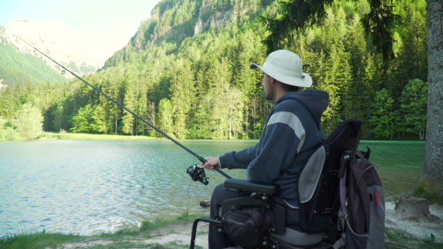 4k-Auflösung-von-behinderten-Fischer-in-einem-elektrischen-Rollstuhl-Angeln-in-schönen-See-in-der-Nähe-von-Wald-und-Berg-im-Rücken
