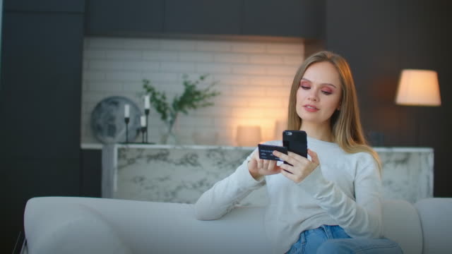 Beautiful-young-female-with-cheerful-expression,-holds-smart-phone-and-credit-card,-banks-online-or-makes-shopping-while-sits-against-cafe-interior.-Payment-and-leisure-concept