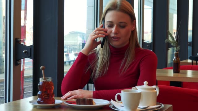 Una-joven-atractiva-mujer-se-sienta-en-una-mesa-con-comida-en-un-café-y-habla-en-un-teléfono-inteligente-con-una-sonrisa