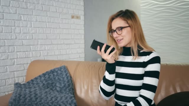 Happy-caucasian-woman-in-glasses-using-voice-recognition-on-phone-to-record-a-message-sitting-on-a-couch-at-home.-Girl-use-mobile-voice-control,-business-woman-in-modern-home