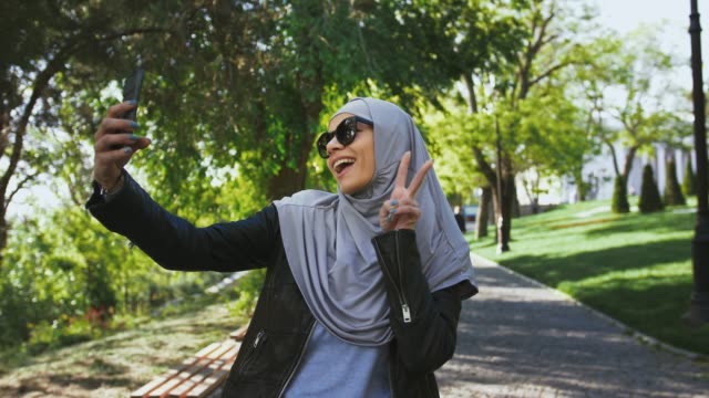 Muslim-woman-in-casual-outfit,-sunglasses-and-hijab.-She-is-smiling,-showing-victory-sign,-taking-selfie-on-her-smartphone,-posing-in-park.-Сlose-up