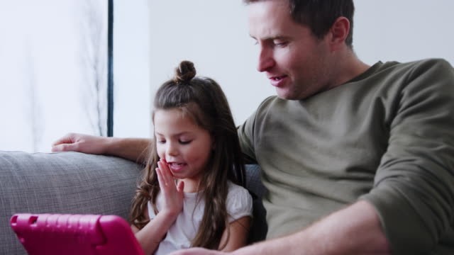 Padre-e-hija-sentados-en-el-sofá-en-casa-jugando-juntos-en-la-tableta-digital-en-el-caso-rosa-en-casa