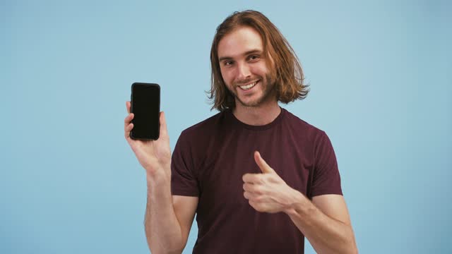 Man-raising-up-his-hand-holding-smartphone-with-black-screen,-pointing-at-it,-showing-thumb-up-and-smiling,-posing-on-blue-background